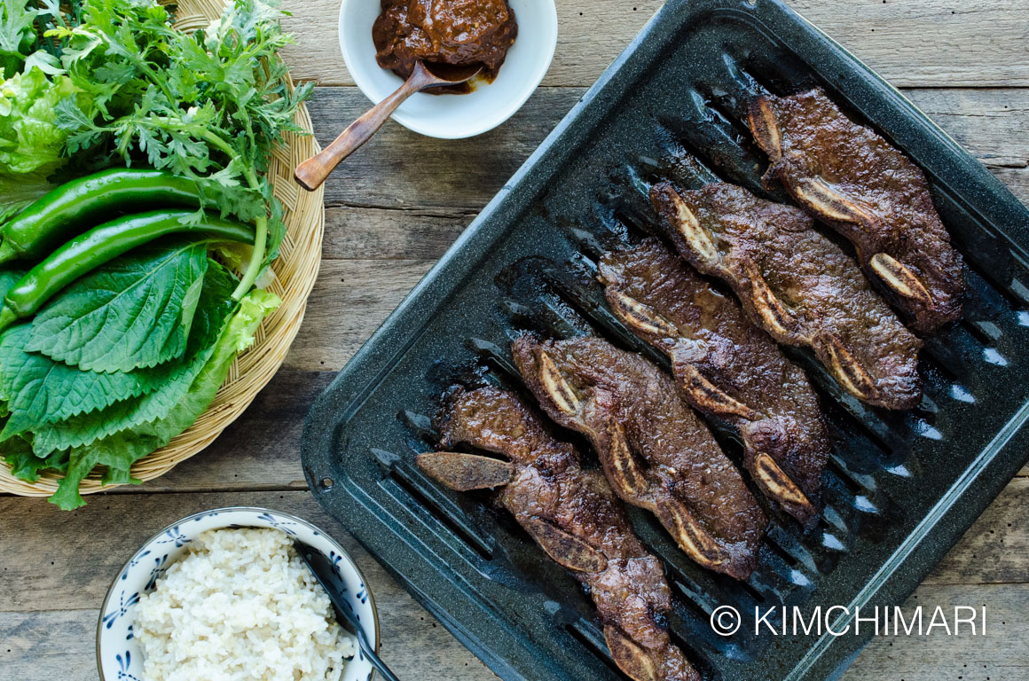 top view of Kalbi Short Ribs on grill pan, ssamjang in a bowl, rice and plate of ssam greens