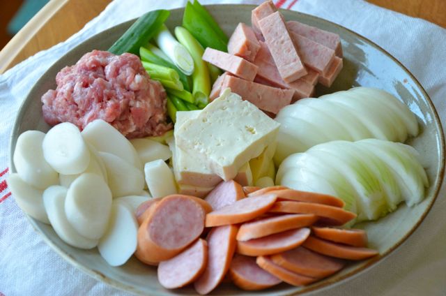 Ingredients for stew plated with rice cake, sausage, onion, spam, green onion, tofu all sliced and ground pork