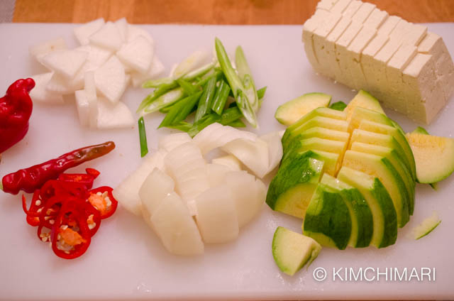 cutting board with cut onion, radish, green onion, tofu, zucchini and peppers for pollock roe stew