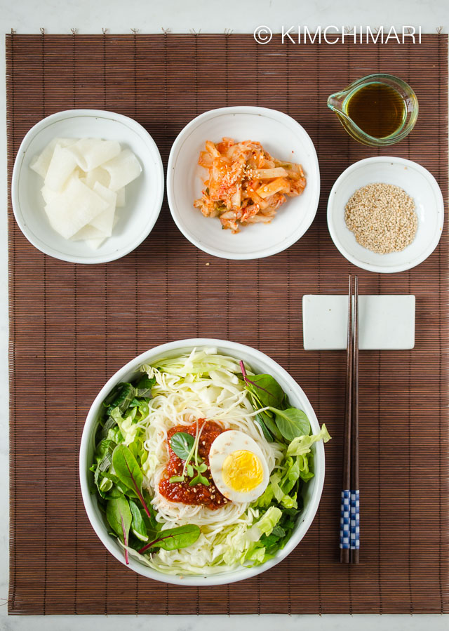 Table Top view of bibim guksu bowl with chopsticks on the side and kimchi and pickled radish as side dishes