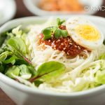 Bowl of bibim guksu served with fresh vegetables , topped with gochujang sauce and egg