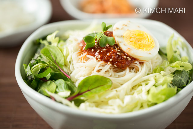 Bowl of bibim guksu served with fresh vegetables , topped with gochujang sauce and egg