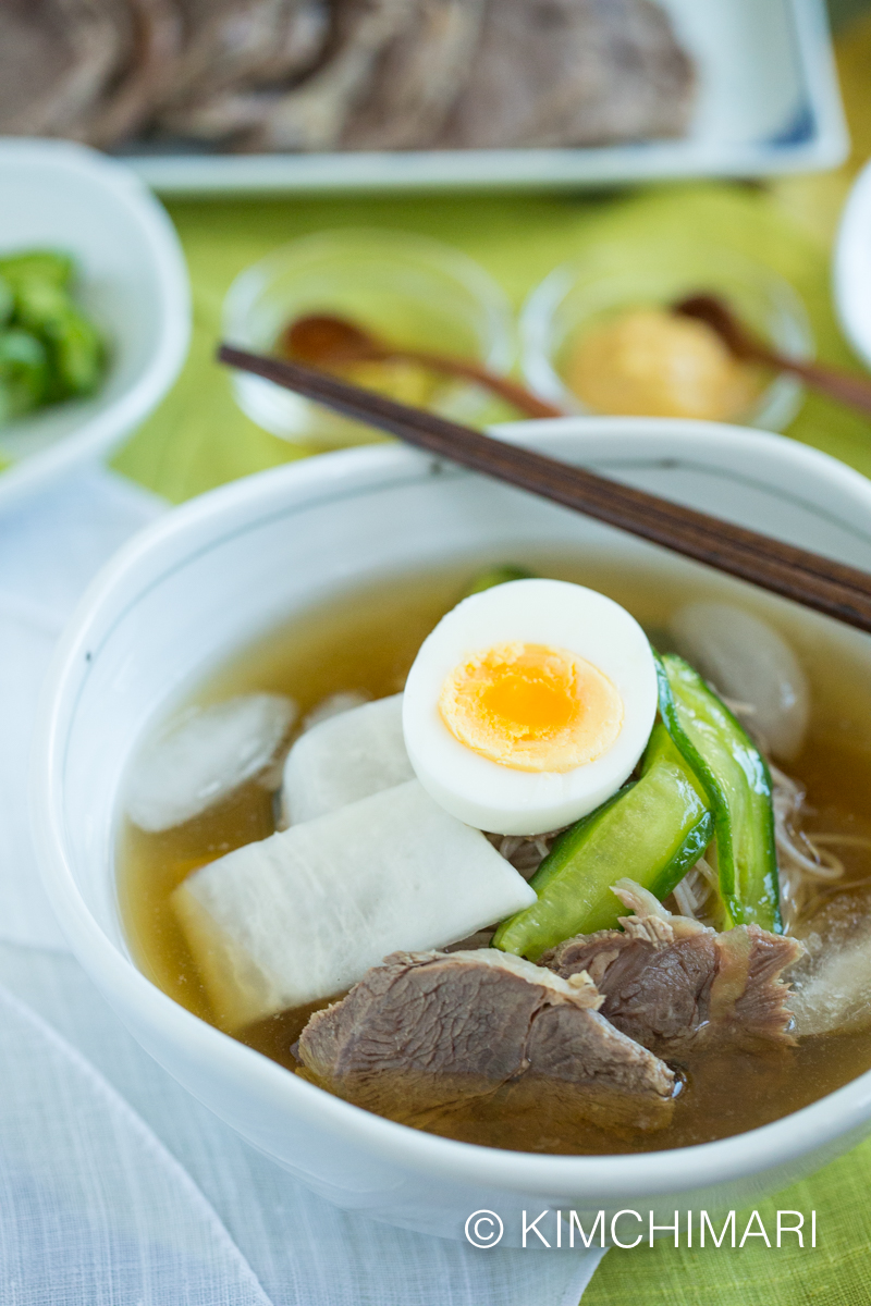 Mul Naengmyeon served in white bowl topped with egg, cucumber, radish and sliced beef