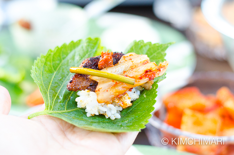 ssam on palm of hand with rice and grilled samgyeopsal, kimchi, ssamjang and garlic scape jangahjji
