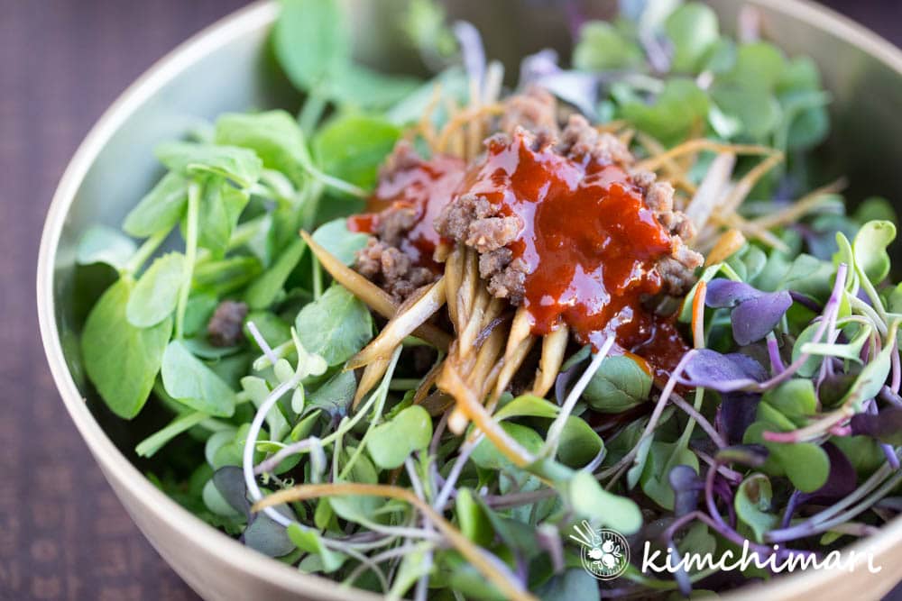 closeup of microgreens on rice, topped with beef and burdock roots and gochujang sauce in a brass bowl