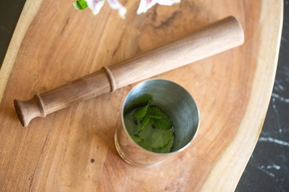 wooden muddler next to cocktail shaker cup with mint leaves in them